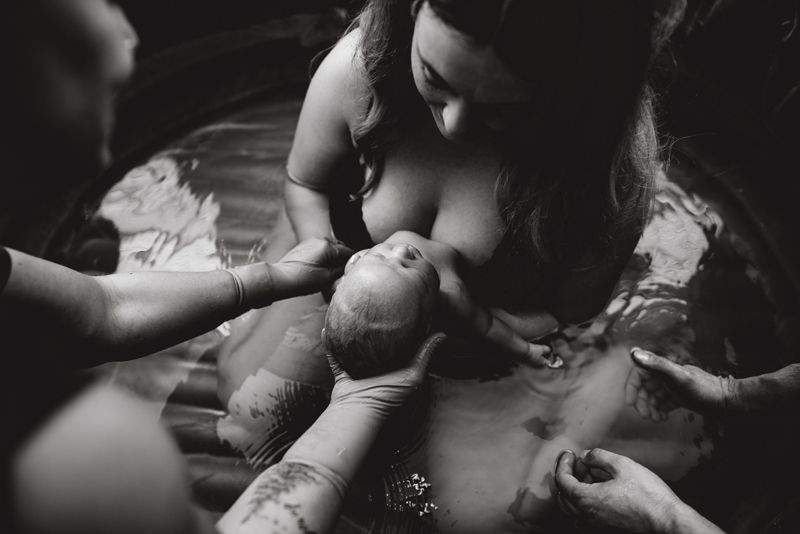 Birth Photography - A woman kneeling in a birthing tub looks down at her newborn baby with admiration