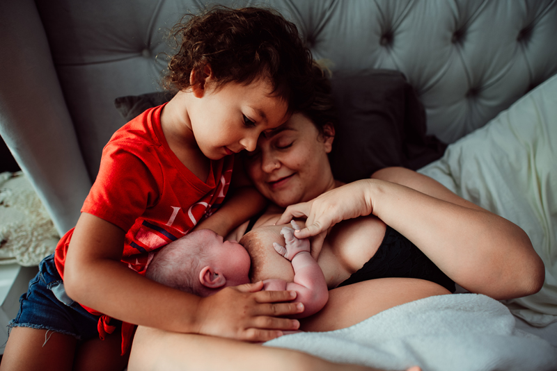 Birth Photography - woman breast feeds newborn baby, her toddler watches and leaning into mom