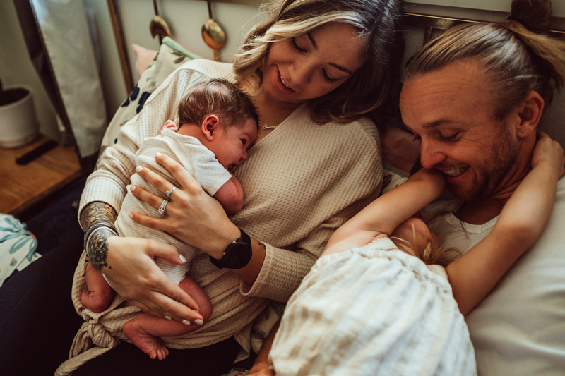 Postpartum Photography - Mom holds onto new baby in bed with dad and their other daughter being playful nearby