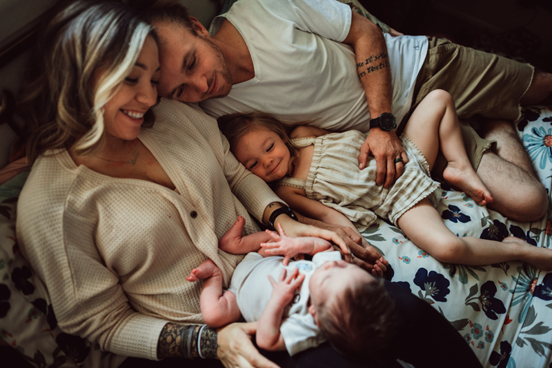 Postpartum Photography - a man and a woman are snuggled together holding their newborn baby and toddler daughter smiling