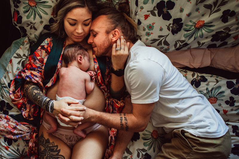 Postpartum Photography - a woman lays in bed with baby on her chest, her husband snuggles them both