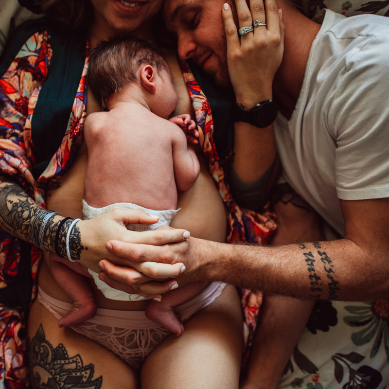 Postpartum Photography - a woman holds her newborn baby on her belly, dad snuggles in holding them both