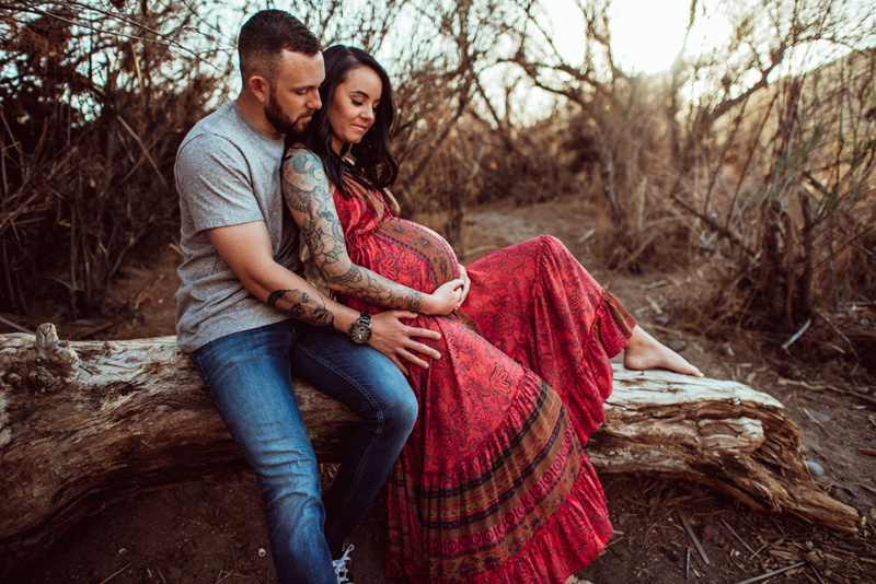 Maternity Photography - woman leans into man as they sit on a large log outdoors, she is expecting
