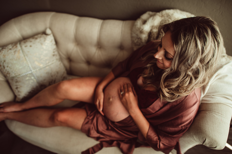 Maternity Photography - a pregnant woman reclines on an elegant couch, she holds her belly happily