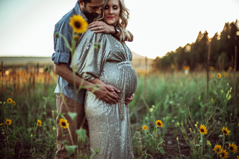 Maternity Photography - a man embraces his wife outdoors in a field of daisies, she is expecting