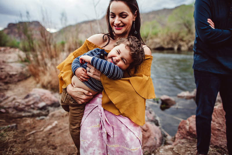 Family Photography - a woman holds her young son, both laughing, they stand near a river