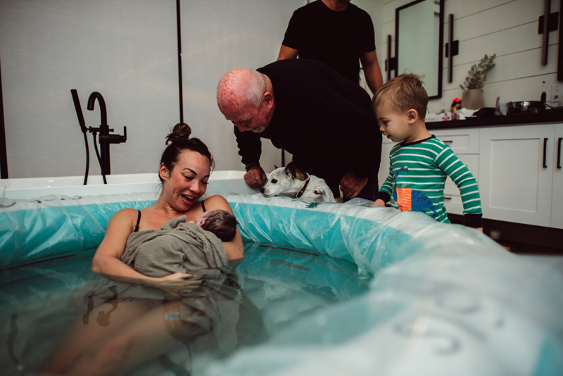 Birth Photography - woman has large smile as she holds her newborn baby. A man, toddler, and little puppy all lean in to the excitement with wonder