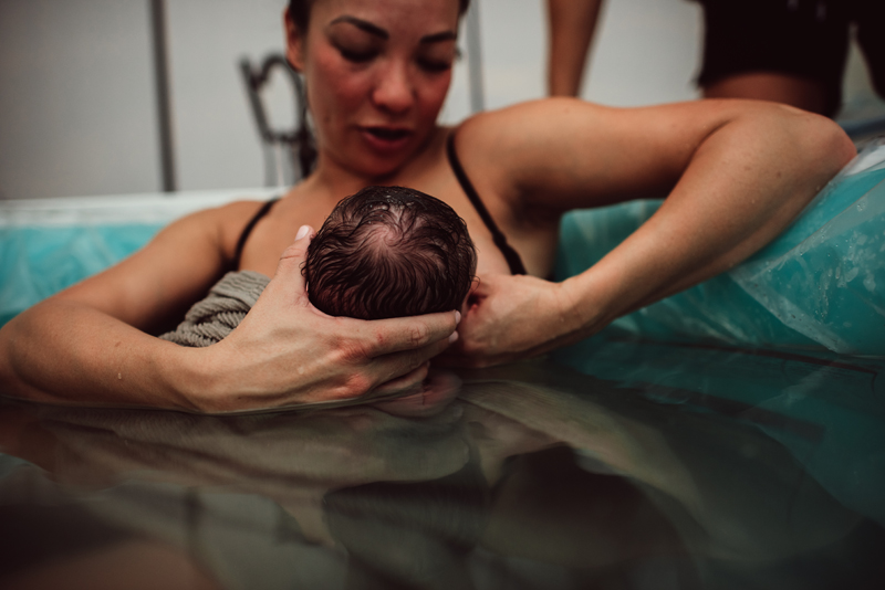 Birth Photography - Mother holds baby close to chest for breastfeeding. She still sits in birthing pool after labor