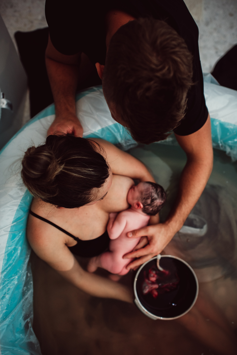 Birth Photography - woman in birthing tub holds baby in her arms as she breastfeeds in her other hand she holds bowl with placenta, dad is near