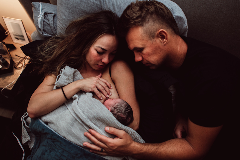 Birth Photography - woman breastfeeds new baby, dad looks on and supports mom