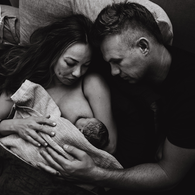 Birth Photography - woman breastfeeds newborn baby, dad leans into mom as they both admire it