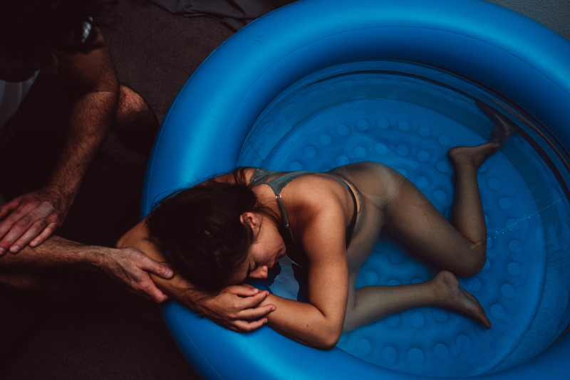 Birth Photography - a woman rests on the side of a birthing pool during labor, her husband soothes her with his hand