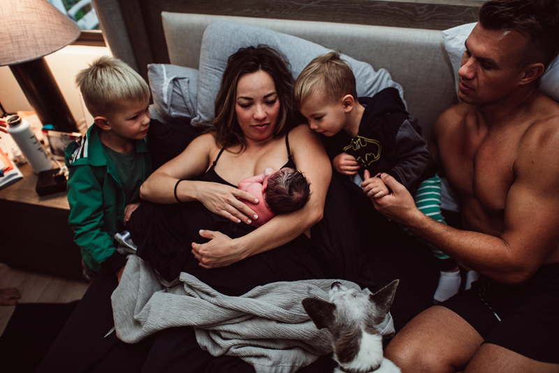 Birth Photography - a woman happily breastfeeds new baby, dad and two little boys lean in to watch