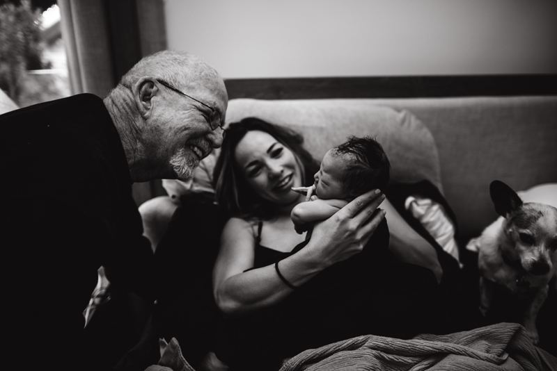 Birth Photography - woman holds up her newborn baby up for grandpa to see, he smiles happily