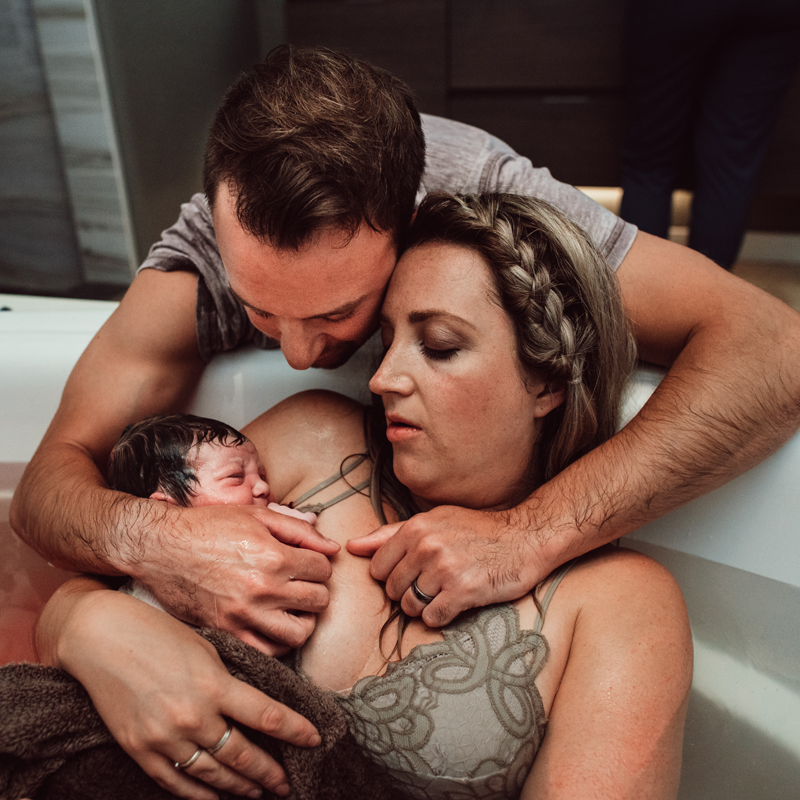 Birth Photography -husband leans over birthing tub to embrace wife and admire new baby