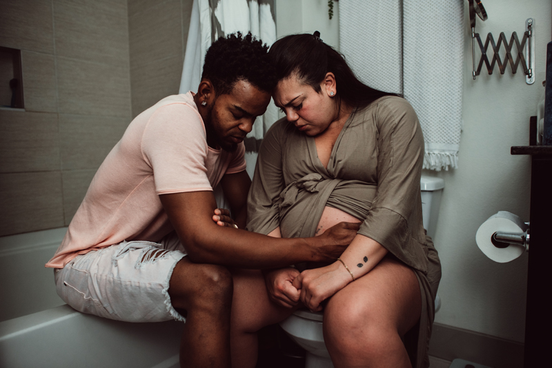 Birth Photography - Man and woman sit in the bathroom leaning into each other. Man holds woman's pregnant belly.