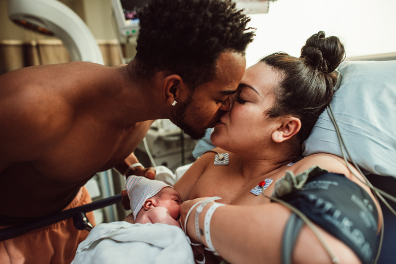 Birth Photography - man gives new mother a kiss on the lips as she holds her newborn baby in the hospital bed