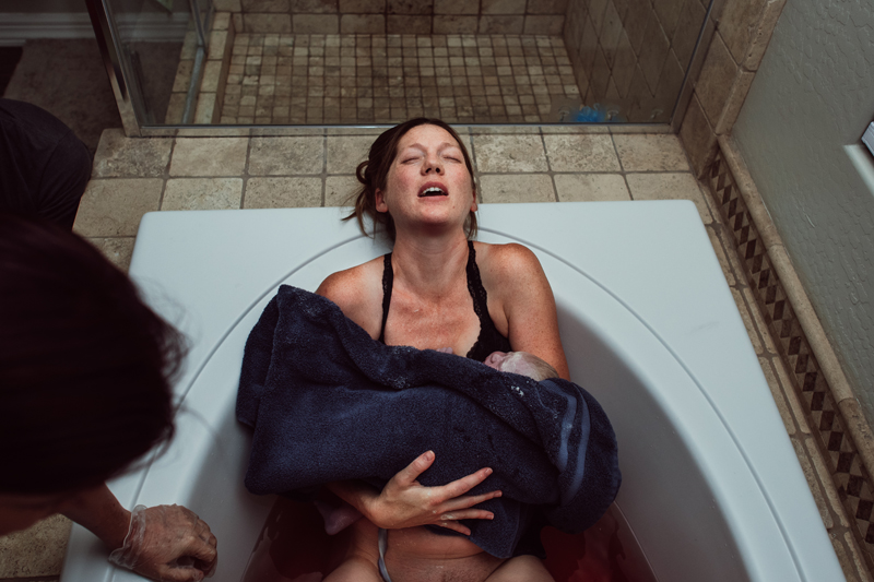 Birth Photography - Woman relaxes as she leans back on bathtub after just giving birth to the newborn baby in her arms