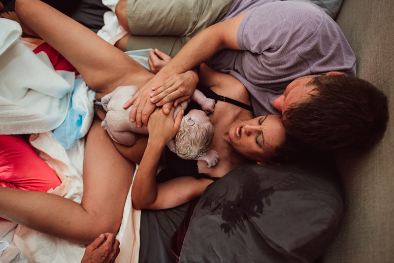 Birth Photography - a woman holds her newborn baby to her chest as she lays in bed. Dad leans into to mom and new baby.