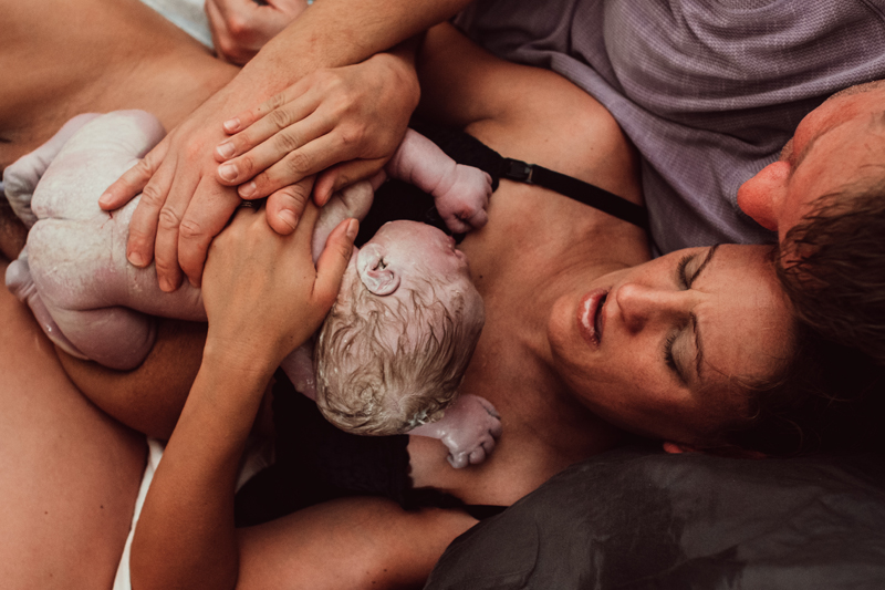 Birth Photography - New mother holds her newborn baby on her chest as she admires it. Dad leans in holding both of them