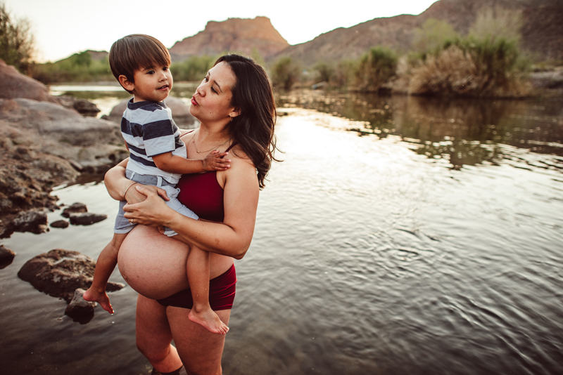 Maternity Photography - A woman in her third trimester holds her young son over her pregnant belly at the river