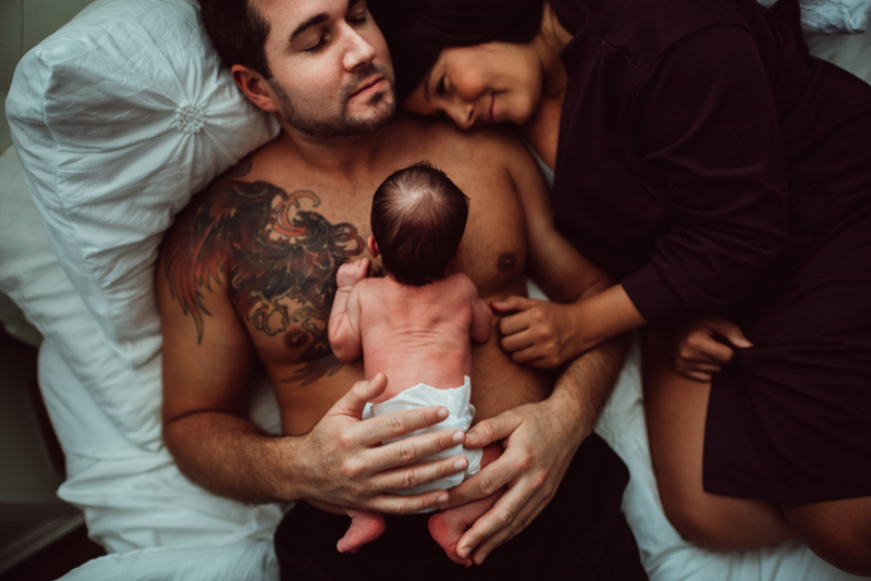 Postpartum Photography - dad lays on bed with newborn child, mom snuggles close to him admiring her family