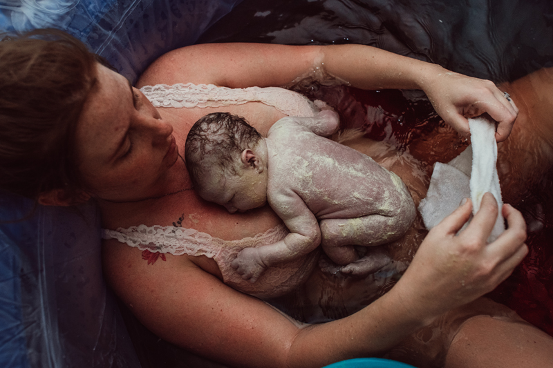 Birth Photography -woman holds just born baby on her chest as she sits in pool from water birth