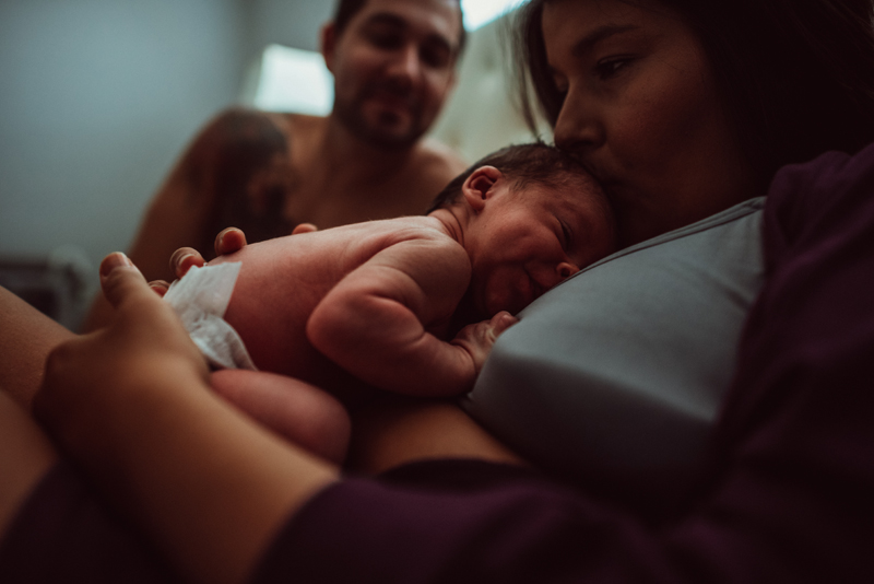 Postpartum Photography - a new mother kisses her baby on the head, dad smiles on in the background