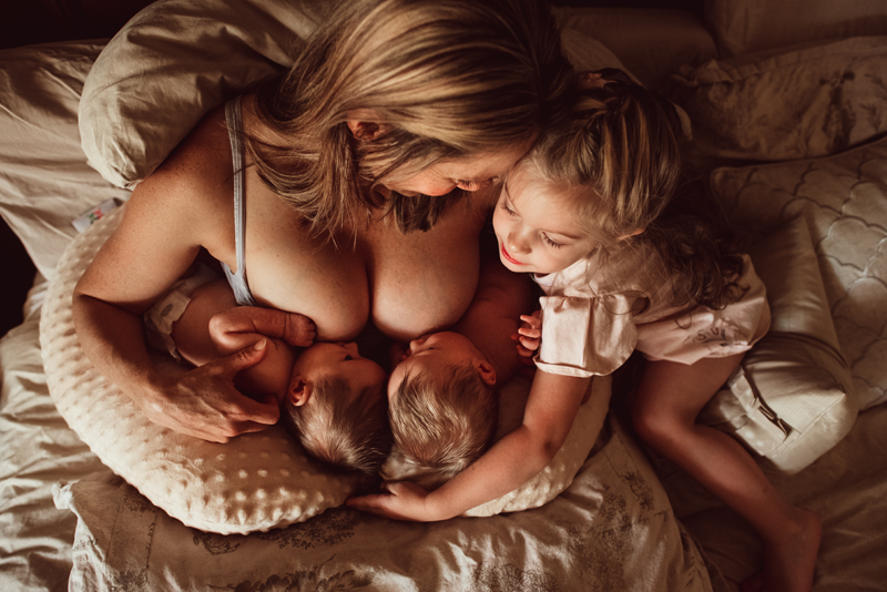 Postpartum Photography - a mother breastfeeds both twins as she sits in bed, her other young daughter admiring her siblings