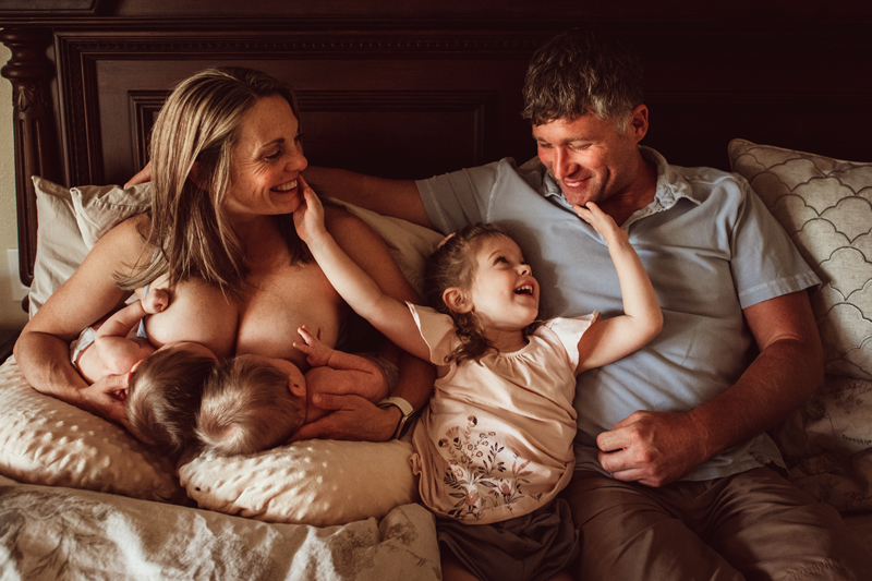 Postpartum Photography - A mother breastfeeds two twins, her other daughter smiles and looks up dad beside them