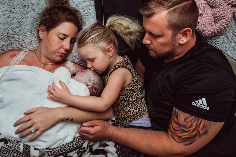 Birth Photography - Family embraces newborn child. Mom holds baby, as sister and dad look on.