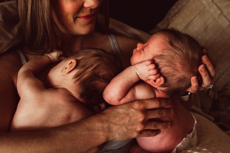 Postpartum Photography - a woman smiles as admires both of her newborn babies close to her chest