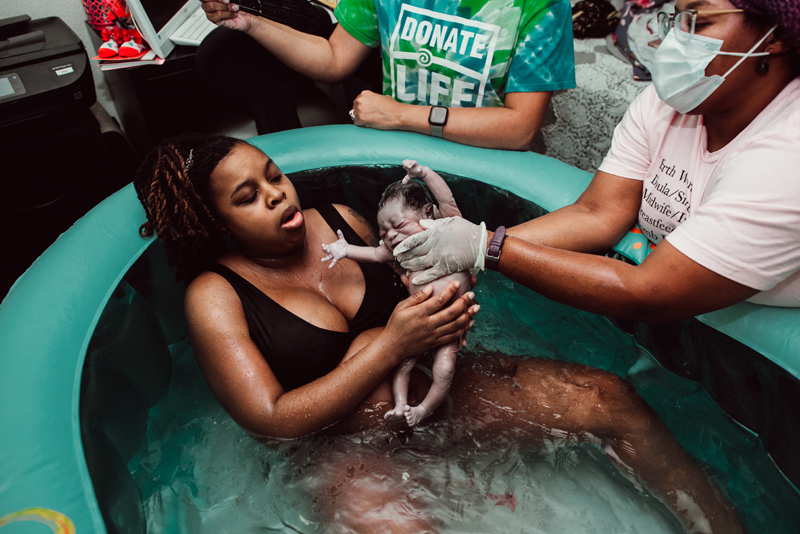Birth Photography - Woman in birthing pool holds newborn baby with doula
