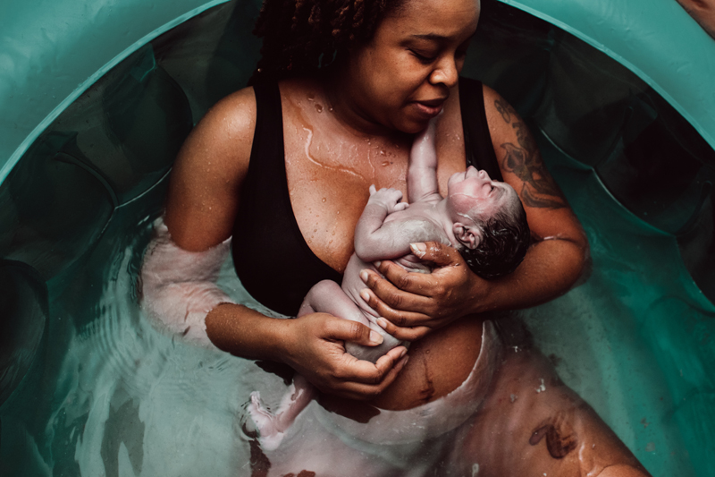 Birth Photography - woman admires newborn baby as she sits in the water from water birth. Baby seemingly looks up at mom