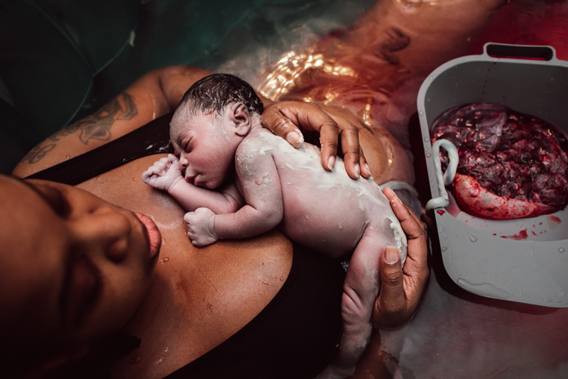 Birth Photography - woman holds newborn baby to chest, placenta sits in bowl nearby, they are in birthing tub