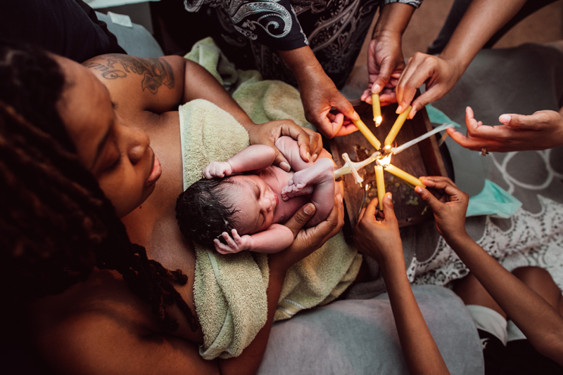 Birth Photography - woman holds her newborn baby, the umbilical cord is clipped and 6 hands hold candle flames to cut the cord