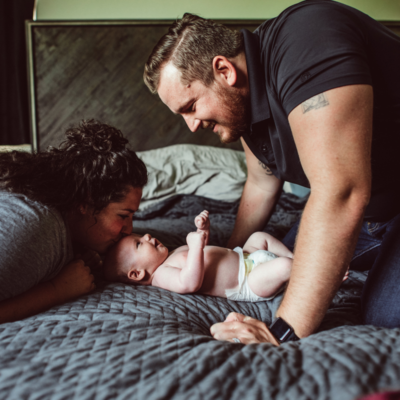 Postpartum Photography - a baby smiles from the bed, dad and mom smiling back at baby