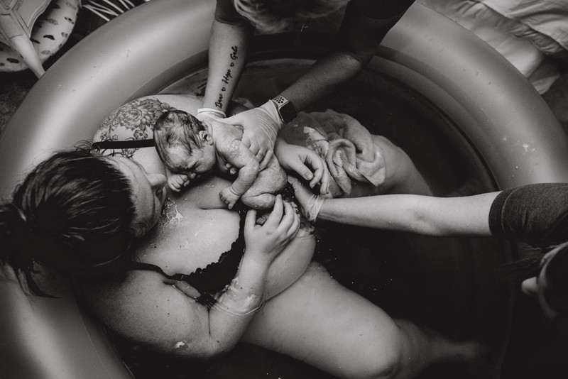 Birth Photography - Mother sits in birthing pool holding newborn baby. The doula and husband's arms reach into to support mom