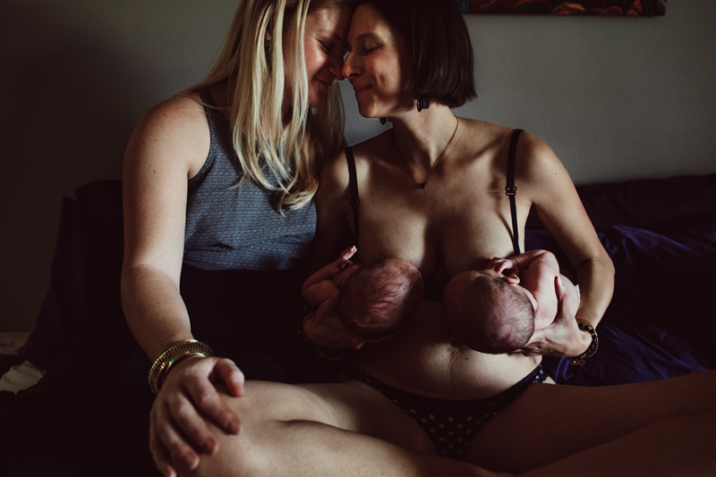 Postpartum Photography - two women look sit close nose to nose, one woman breastfeeds both twins