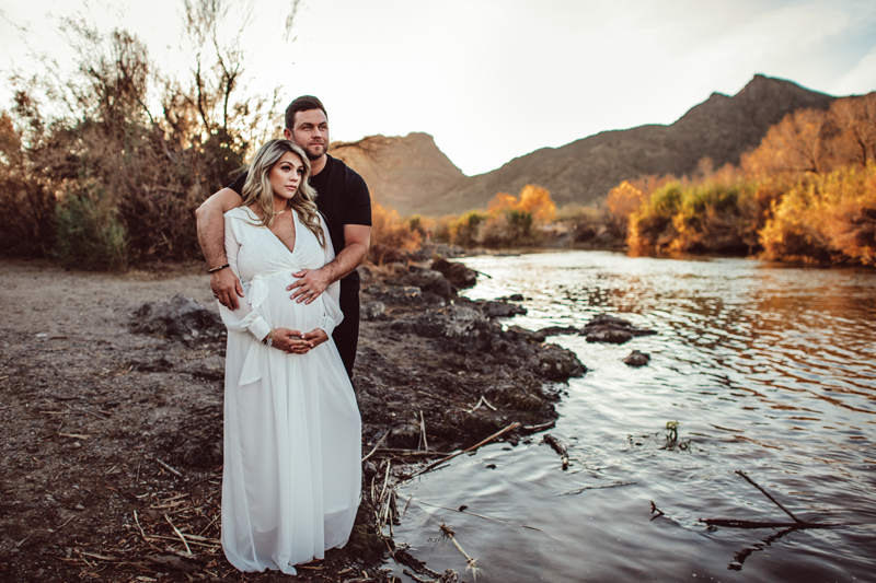 Maternity Photography - man embraces wife, she is in a white flowing dress pregnant, they stand near the river's bank..