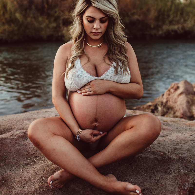Maternity Photography - a woman sits near a river bank, she is crossed legged and holds her pregnant belly