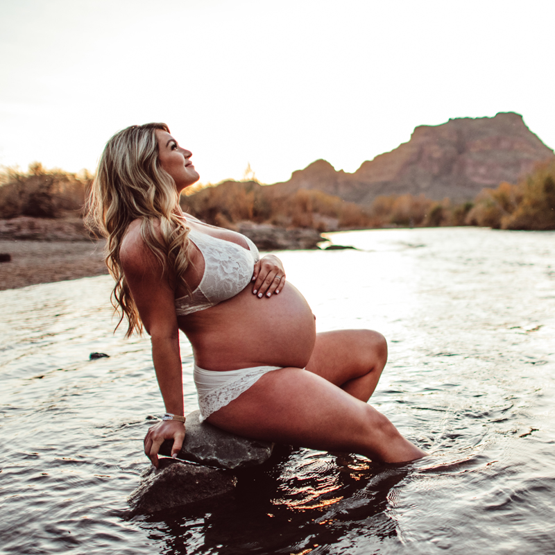 Maternity Photography - a pregnant woman sits on a rock in a quiet river happily