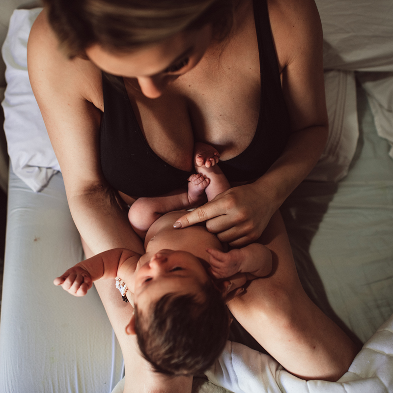 Postpartum Photography - a woman sits in bed holding onto her newborn baby