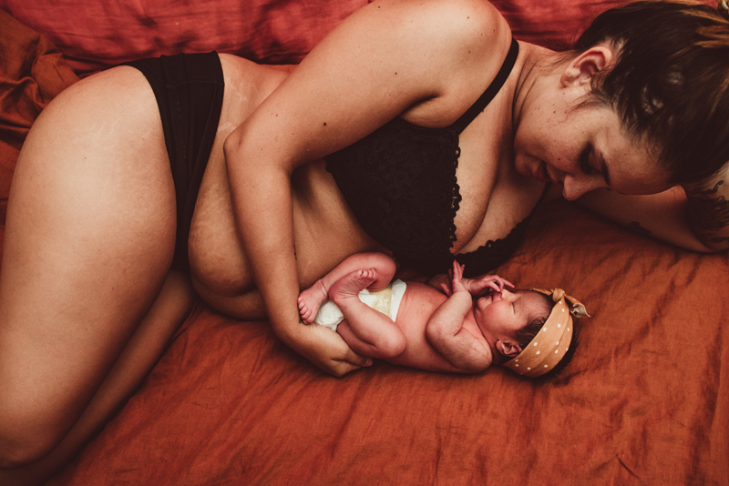 Postpartum Photography - a woman lays on her bed as she admires and loves her newborn baby daughter laying bedside her