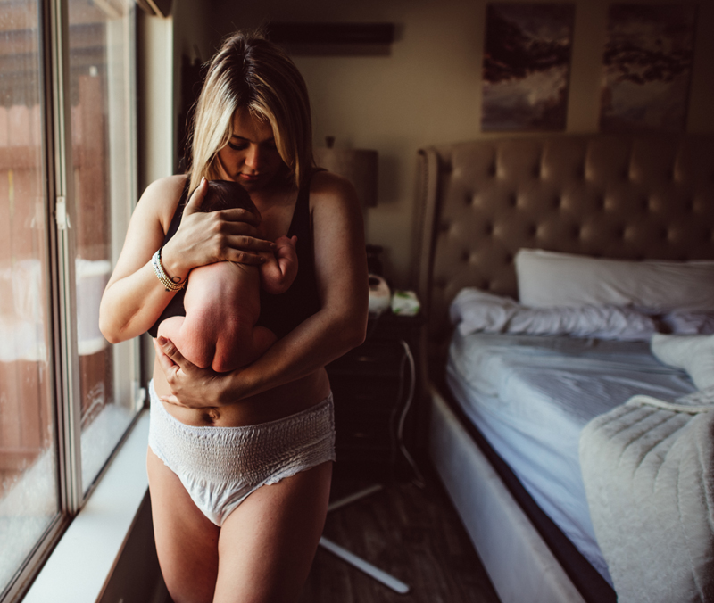 Postpartum Photography - a woman stands in her bedroom holding her newborn baby tightly