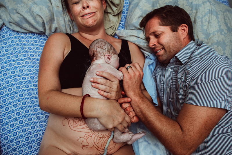 Birth Photography - woman lays in hospital bed holding newborn baby, her husband next to her, both crying