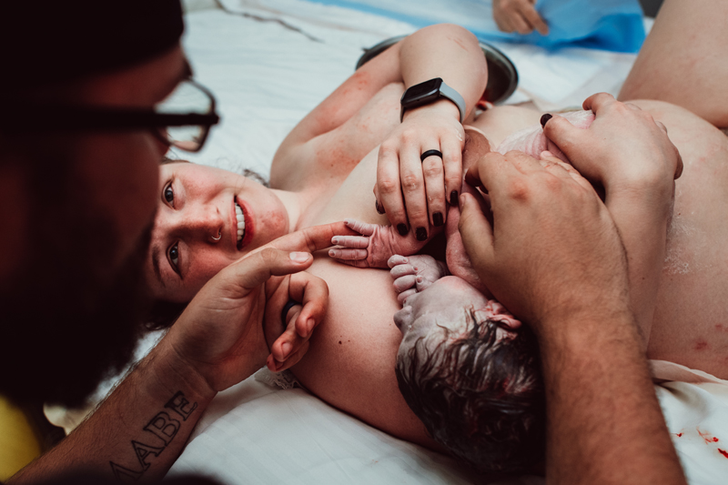 Birth Photography - New mom lays in bed happily smiling having just delivered the newborn child in her arms