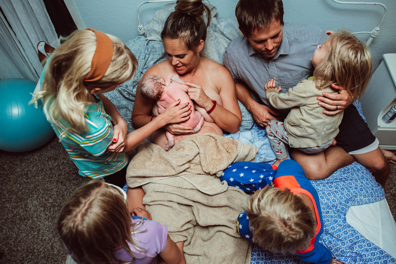 Birth Photography - mom lays in bed happy as newborn baby latches on to breastfeed. Dad is close with four daughters