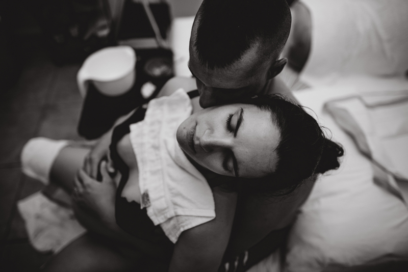 Birth Photography - A woman concentrates as she holds her pregnant belly. Her husband embraces soon-to-be mom from behind