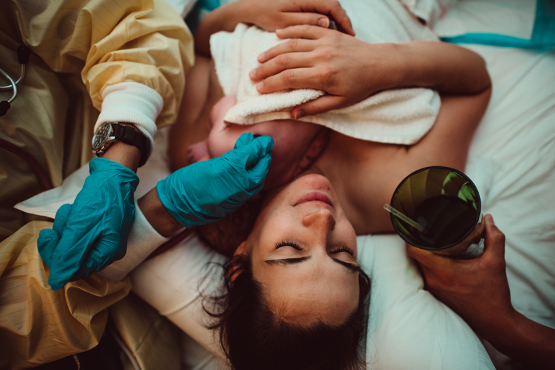 Birth Photography - a woman looks full with joy holding her newborn baby as she lays with her eyes closed, dad provides her a glass for water nearby